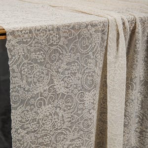 May include: A cream-colored lace tablecloth with a floral and swirl pattern. The delicate fabric drapes over a table, showcasing the intricate design. The lace is a neutral color, suitable for various events.