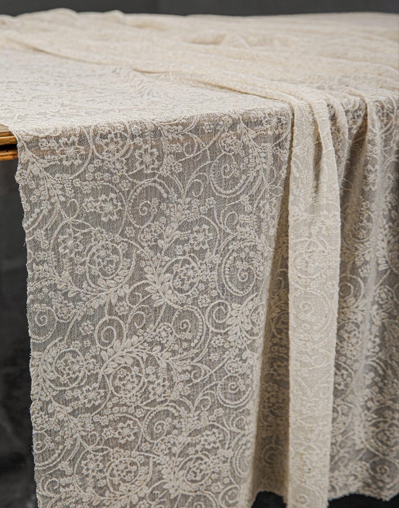 May include: A cream-colored lace tablecloth with a floral and swirl pattern. The delicate fabric drapes over a table, showcasing the intricate design. The lace is a neutral color, suitable for various events.