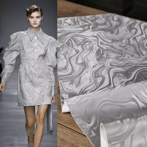 May include: A light grey dress with a marbled pattern, featuring a collared neckline and button-down front. The dress has long sleeves with puffed shoulders. A roll of matching marbled fabric is displayed next to the dress.