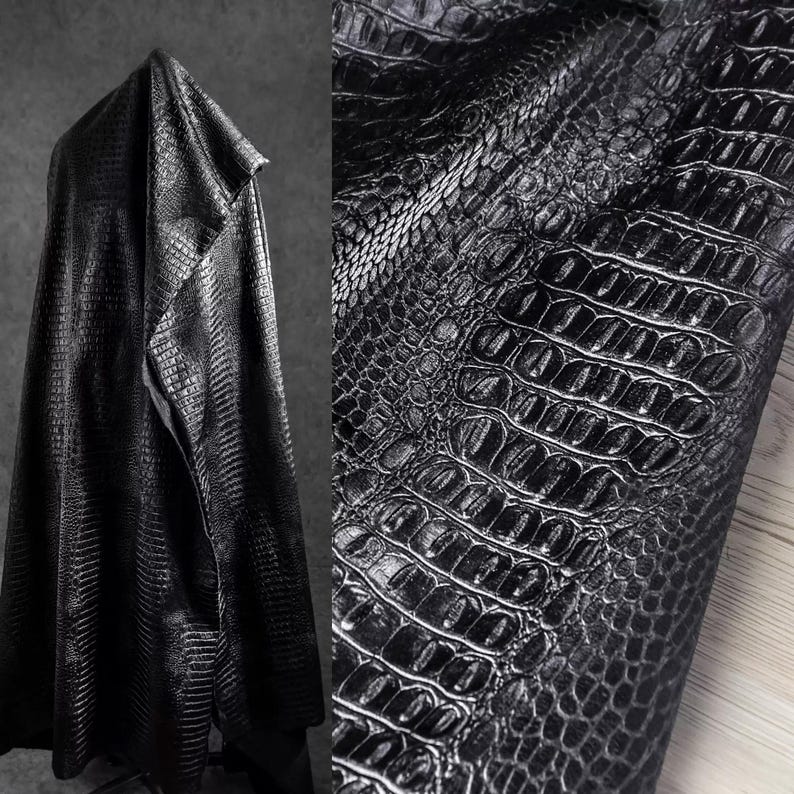 May include: Black faux leather fabric with a crocodile skin texture. The fabric is draped over a hanger, with a close-up view of the detailed pattern. The material has a glossy finish, reflecting light.