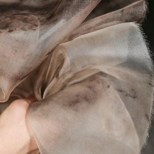 May include: Close-up of a sheer, flowing fabric with a watercolor-like pattern. The fabric is primarily a light beige color with darker brown and gray accents. The edges of the fabric have a delicate, fringed texture.