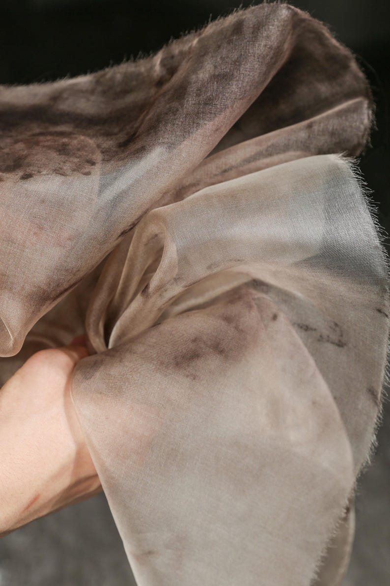 May include: Close-up of a sheer, flowing fabric with a watercolor-like pattern. The fabric is primarily a light beige color with darker brown and gray accents. The edges of the fabric have a delicate, fringed texture.