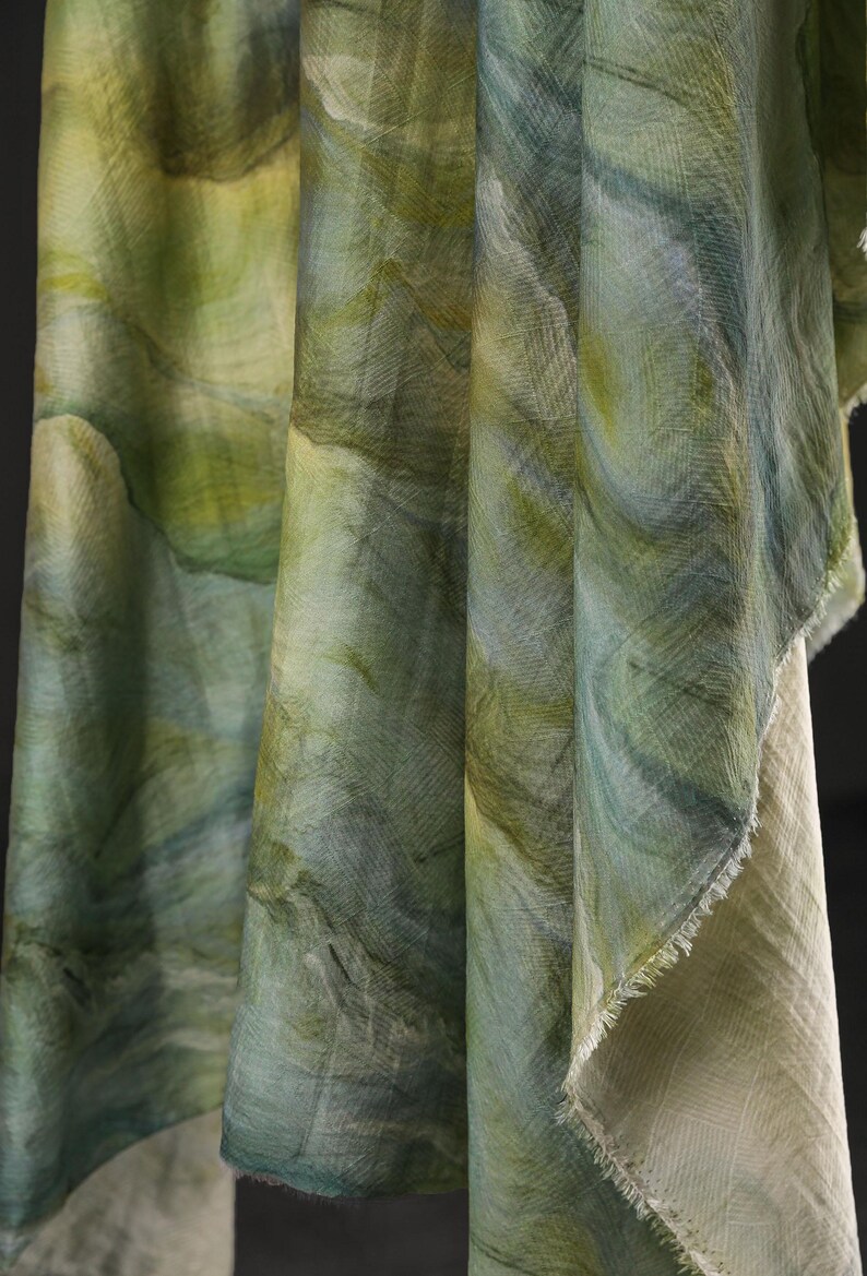 Puede incluir: Primer plano de tela drapeada con un patr&oacute;n similar a la acuarela en tonos verdes y azules. La tela tiene un aspecto texturizado con pinceladas visibles y un borde sin rematar. El efecto general es suave y fluido, lo que sugiere un textil.