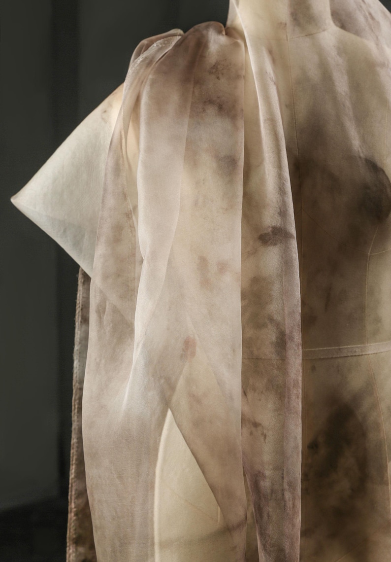 May include: A sheer, cream-colored fabric with brown, mottled patterns draped over a dress form. The fabric is lightweight and appears to be silk or a similar material. The design suggests a focus on textile art or fashion.