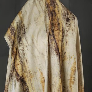 May include: Cream-colored fabric with a marbled pattern in shades of brown and yellow. The fabric is draped, creating folds and shadows that highlight the texture and color variations. The overall effect is organic and earthy.