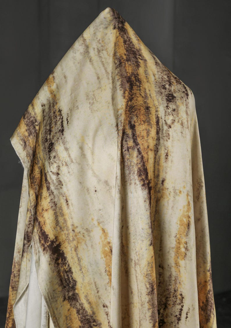 May include: Cream-colored fabric with a marbled pattern in shades of brown and yellow. The fabric is draped, creating folds and shadows that highlight the texture and color variations. The overall effect is organic and earthy.