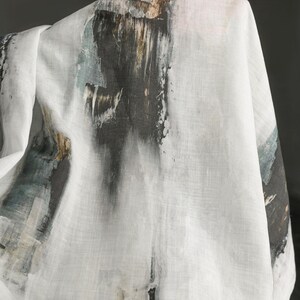 May include: White fabric with an abstract design featuring dark grey, brown, and teal brushstrokes. The fabric appears to be draped, showcasing the artistic pattern. The design resembles a watercolour painting.