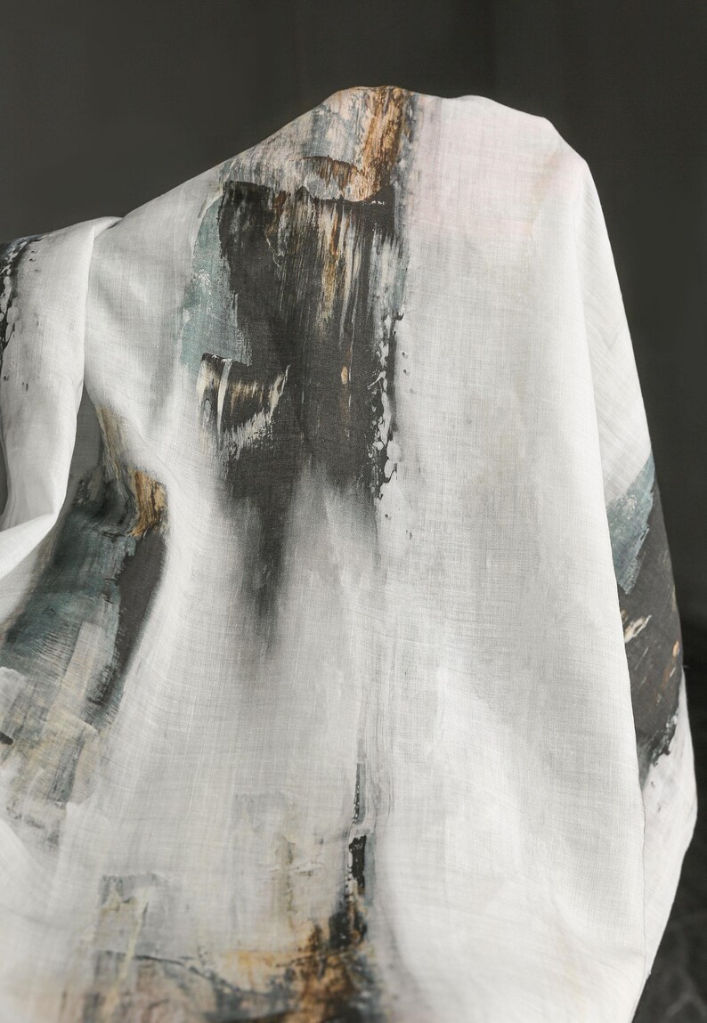 May include: White fabric with an abstract design featuring dark grey, brown, and teal brushstrokes. The fabric appears to be draped, showcasing the artistic pattern. The design resembles a watercolour painting.