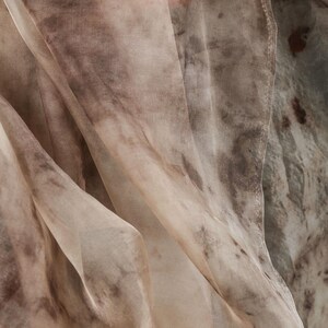May include: Close-up of a flowing fabric with a watercolor-like pattern. The fabric features a blend of beige, brown, and gray tones, creating a soft, textured appearance. The material drapes in gentle folds, suggesting a lightweight textile.