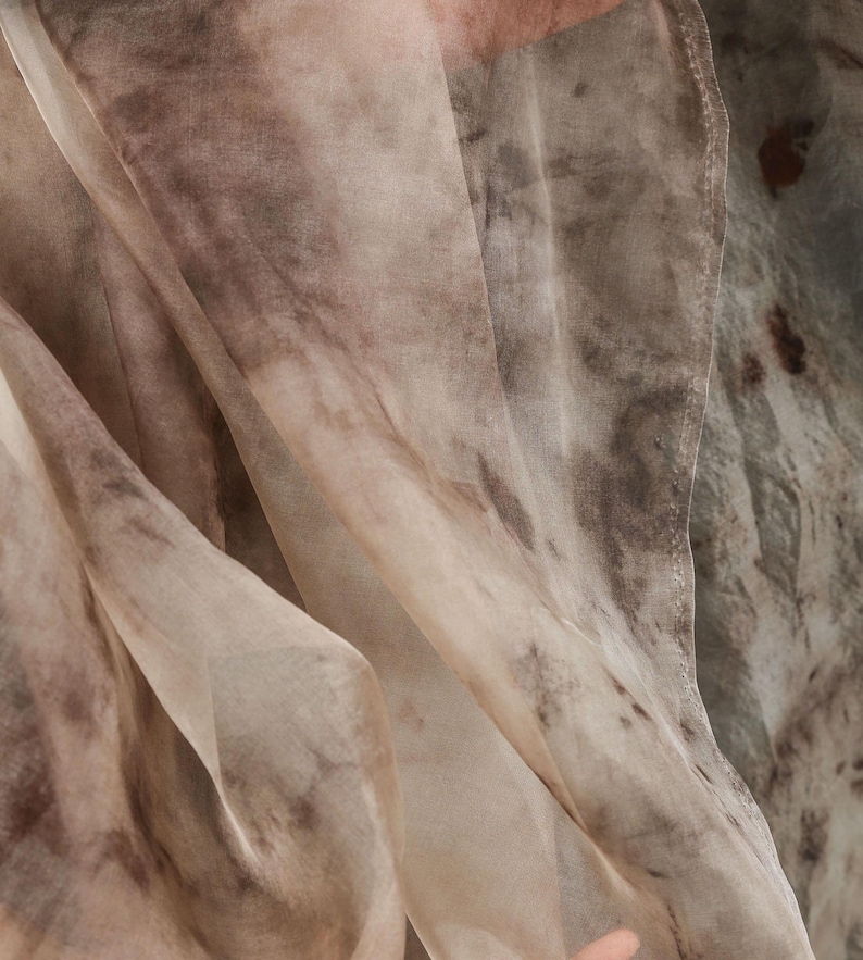 May include: Close-up of a flowing fabric with a watercolor-like pattern. The fabric features a blend of beige, brown, and gray tones, creating a soft, textured appearance. The material drapes in gentle folds, suggesting a lightweight textile.