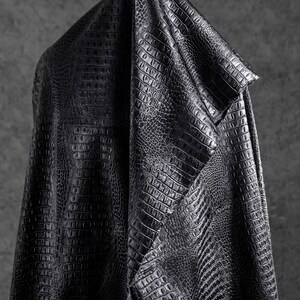 May include: A draped piece of black faux leather fabric with a crocodile skin texture. The material is folded over, showcasing the detailed pattern and the way it catches the light, creating shadows and highlights.
