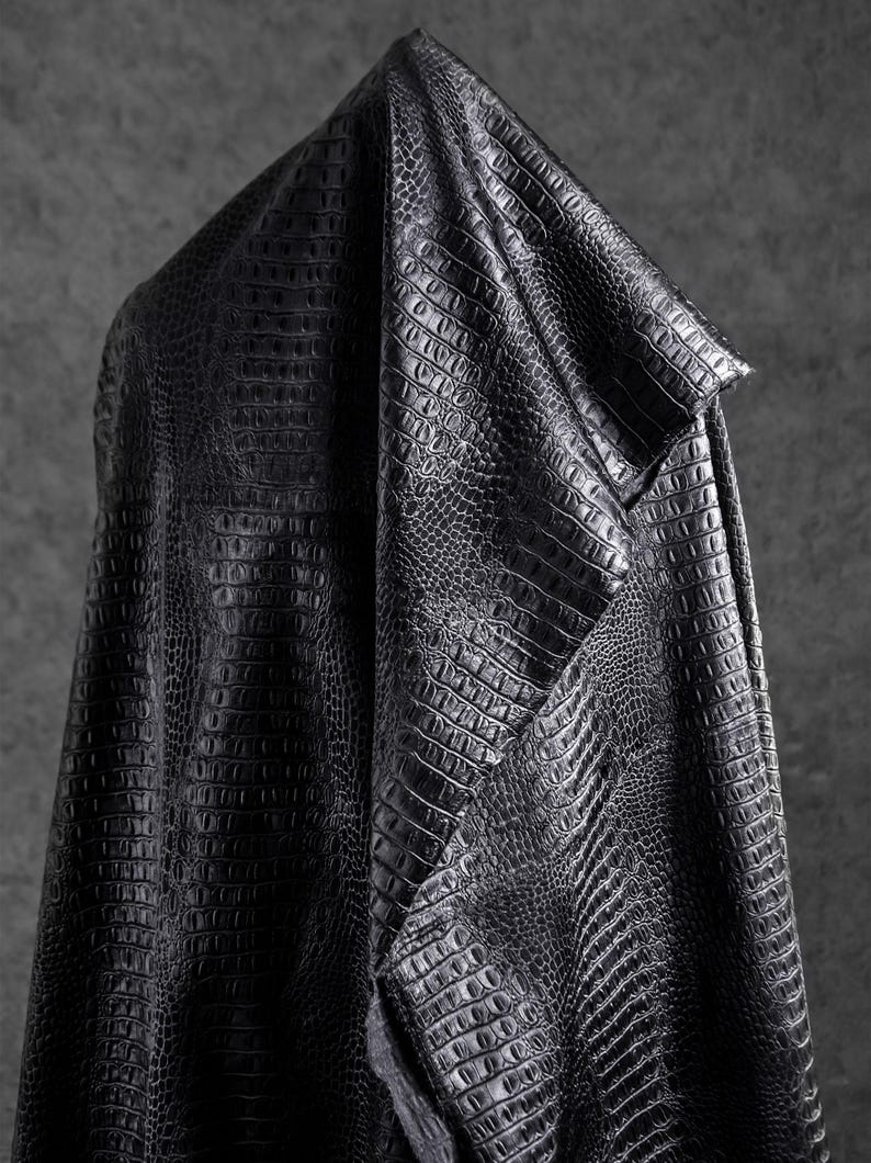 May include: A draped piece of black faux leather fabric with a crocodile skin texture. The material is folded over, showcasing the detailed pattern and the way it catches the light, creating shadows and highlights.
