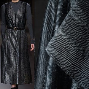 May include: A long, black dress with a V-neck and a matching belt. The dress has a textured, ribbed pattern. The model is wearing a long-sleeved, dark gray top underneath. The fabric has a subtle sheen.
