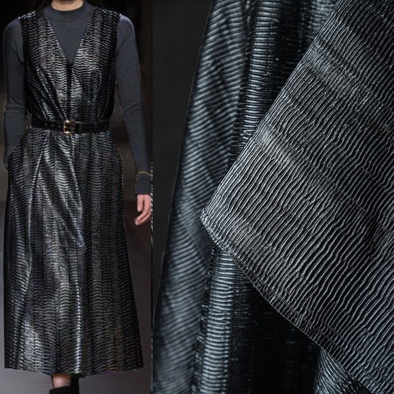 May include: A long, black dress with a V-neck and a matching belt. The dress has a textured, ribbed pattern. The model is wearing a long-sleeved, dark gray top underneath. The fabric has a subtle sheen.