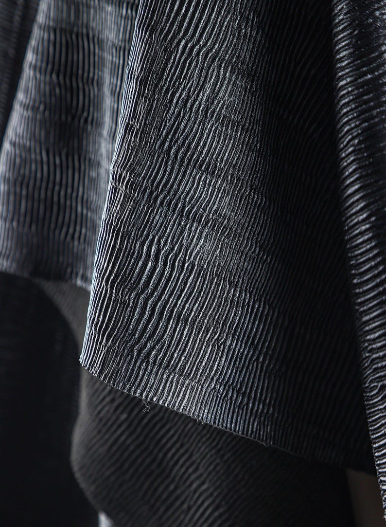 May include: Close-up of a textured, dark gray fabric with a ribbed pattern. The fabric appears to be draped, with folds and shadows adding depth. The surface has a subtle sheen, enhancing the visual texture.