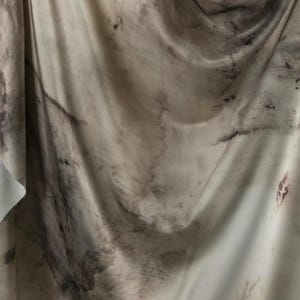 May include: A draped fabric with a marbled appearance. The material is primarily off-white with dark brown and black organic patterns. The fabric's folds create shadows and highlights, enhancing the texture and visual depth. The overall aesthetic suggests a natural, artistic textile.