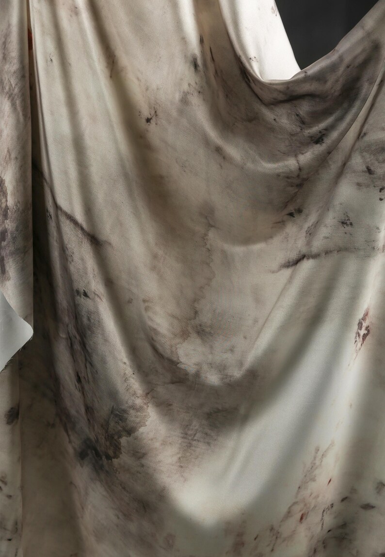 May include: A draped fabric with a marbled appearance. The material is primarily off-white with dark brown and black organic patterns. The fabric's folds create shadows and highlights, enhancing the texture and visual depth. The overall aesthetic suggests a natural, artistic textile.