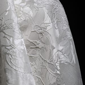K&ouml;nnte beinhalten: Nahaufnahme eines wei&szlig;en Stoffes mit floralem Muster. Der Stoff hat eine strukturierte Oberfl&auml;che mit erhabenen floralen Designs. Die Falten des Stoffes sind sichtbar und erzeugen Schatten und Glanzlichter. Der Hintergrund ist schwarz.