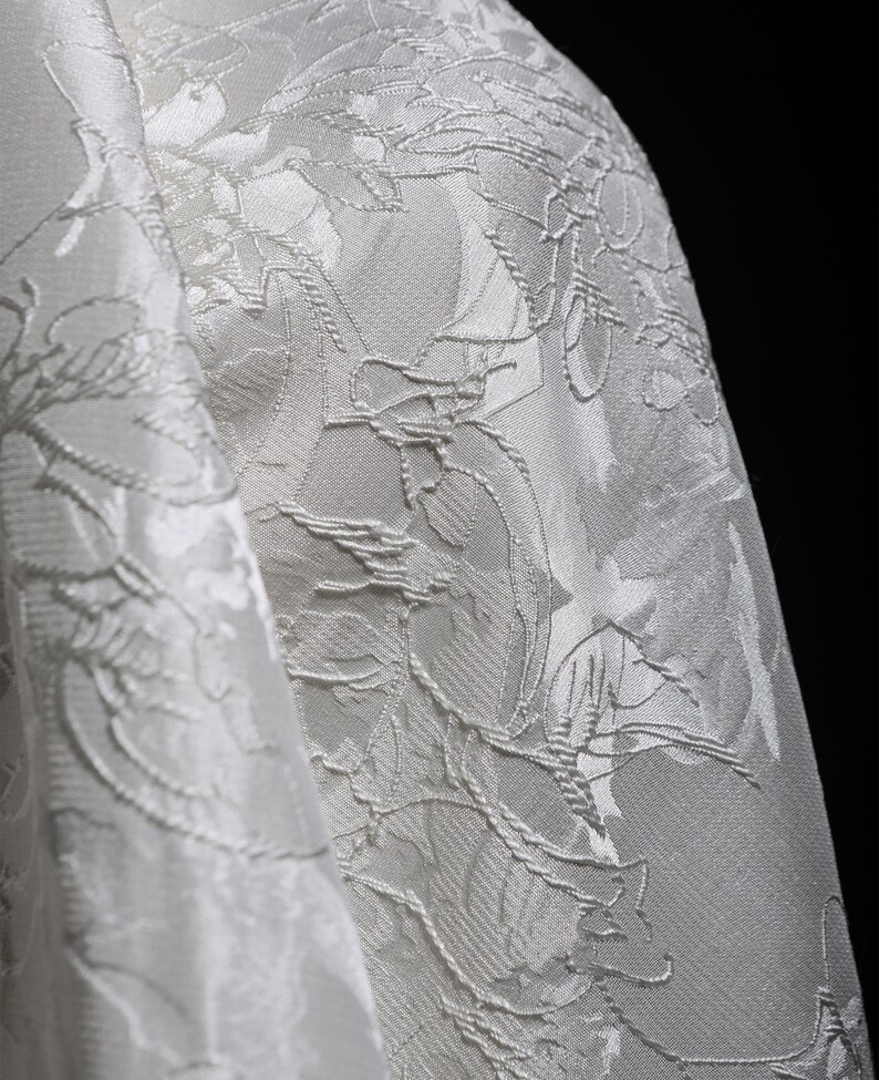 K&ouml;nnte beinhalten: Nahaufnahme eines wei&szlig;en Stoffes mit floralem Muster. Der Stoff hat eine strukturierte Oberfl&auml;che mit erhabenen floralen Designs. Die Falten des Stoffes sind sichtbar und erzeugen Schatten und Glanzlichter. Der Hintergrund ist schwarz.