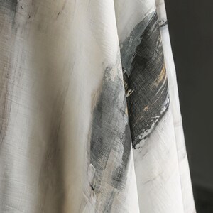 May include: Close-up of a white fabric featuring abstract brushstroke designs in grey, black, and gold. The fabric has a textured appearance, with visible weave details. The overall aesthetic is artistic and modern.