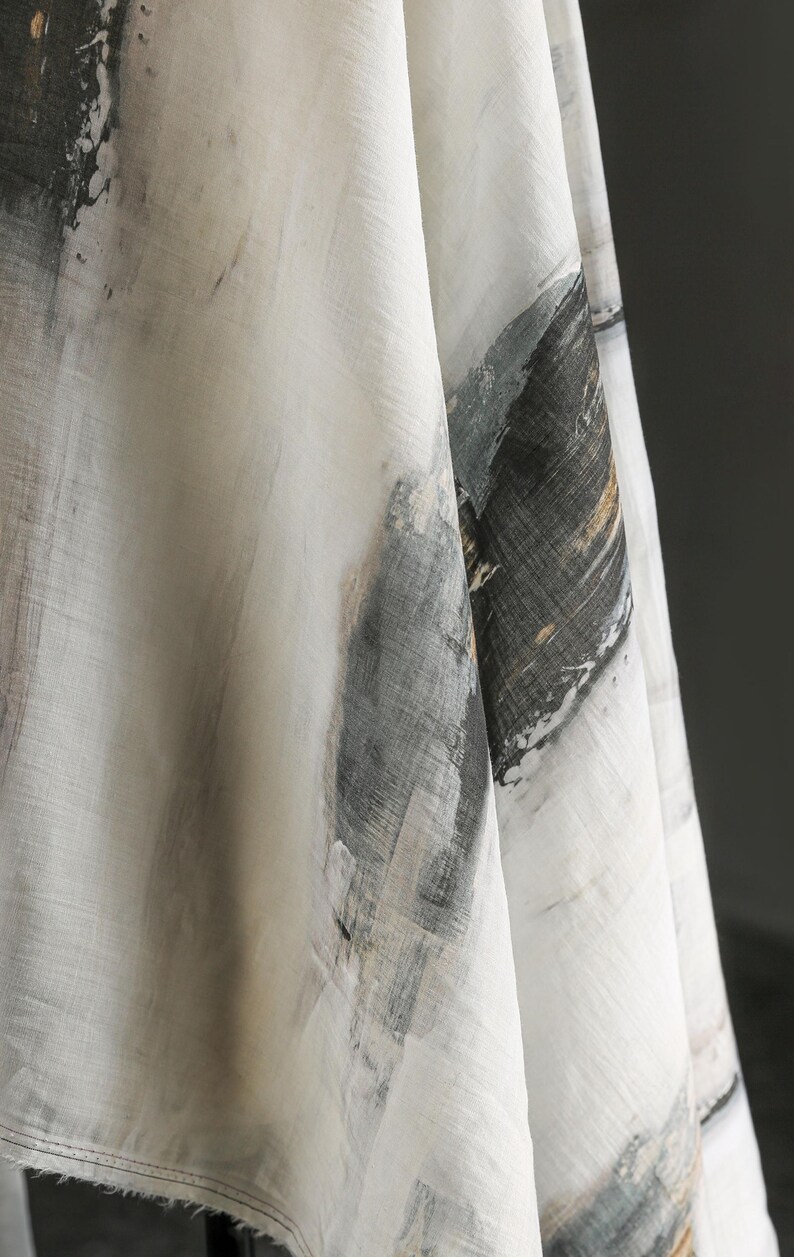 May include: Close-up of a white fabric featuring abstract brushstroke designs in grey, black, and gold. The fabric has a textured appearance, with visible weave details. The overall aesthetic is artistic and modern.