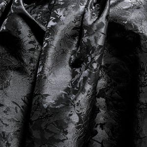 K&ouml;nnte beinhalten: Nahaufnahme eines schwarzen Stoffes mit floralem Muster. Der Stoff ist drapiert und erzeugt Falten und Schatten, die das komplizierte Design hervorheben. Das Material hat einen subtilen Glanz, der der dunklen Farbe Tiefe verleiht.