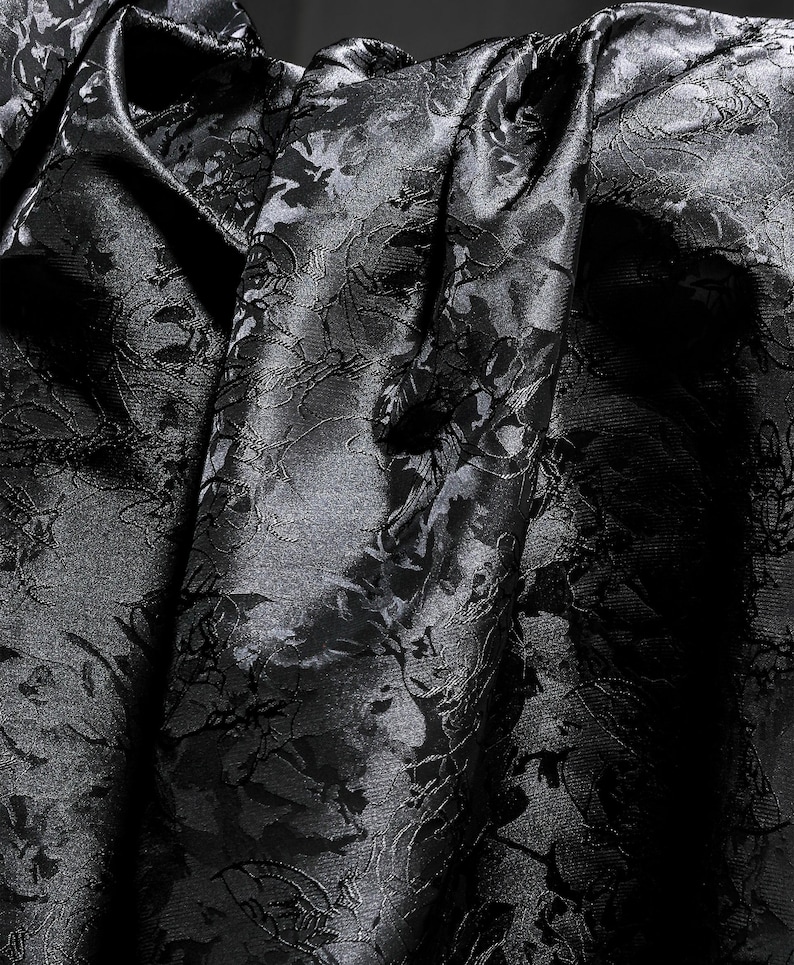 K&ouml;nnte beinhalten: Nahaufnahme eines schwarzen Stoffes mit floralem Muster. Der Stoff ist drapiert und erzeugt Falten und Schatten, die das komplizierte Design hervorheben. Das Material hat einen subtilen Glanz, der der dunklen Farbe Tiefe verleiht.