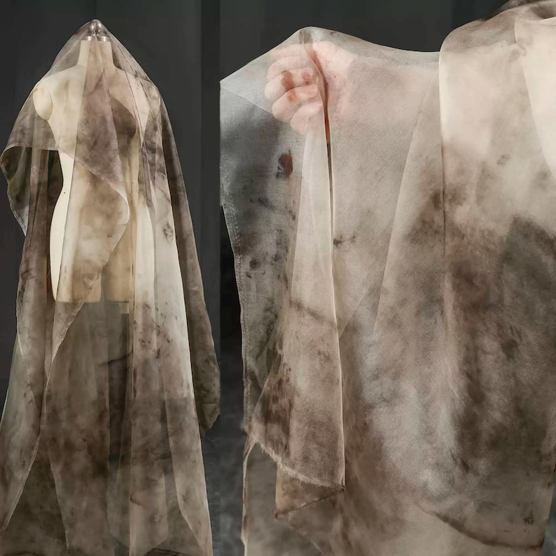 May include: Sheer fabric with a watercolor-like pattern in shades of brown and beige. The fabric is draped over a mannequin and held up by a hand, showcasing its delicate texture and flow. The fabric appears to be lightweight and suitable for various craft projects.