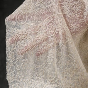 May include: Close-up of a cream-colored lace fabric with an intricate floral and scroll design. The delicate lace is draped, revealing the texture and pattern. The fabric is likely used for apparel or home decor.