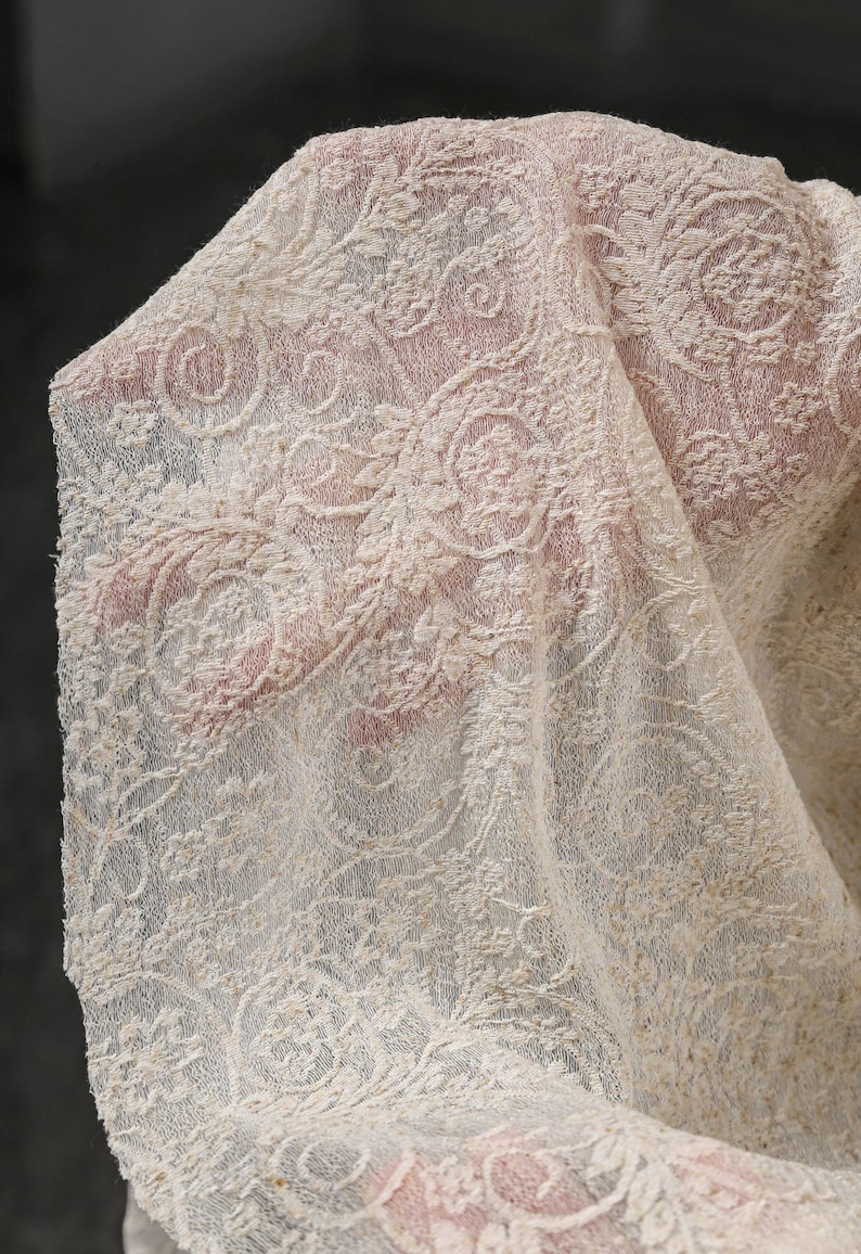 May include: Close-up of a cream-colored lace fabric with an intricate floral and scroll design. The delicate lace is draped, revealing the texture and pattern. The fabric is likely used for apparel or home decor.