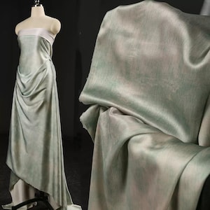 May include: A draped, floor-length gown on a mannequin, featuring a soft, flowing fabric in shades of pale green and white. The dress has a strapless bodice with a white band and a draped skirt. The fabric appears to have a subtle, watercolor-like effect.