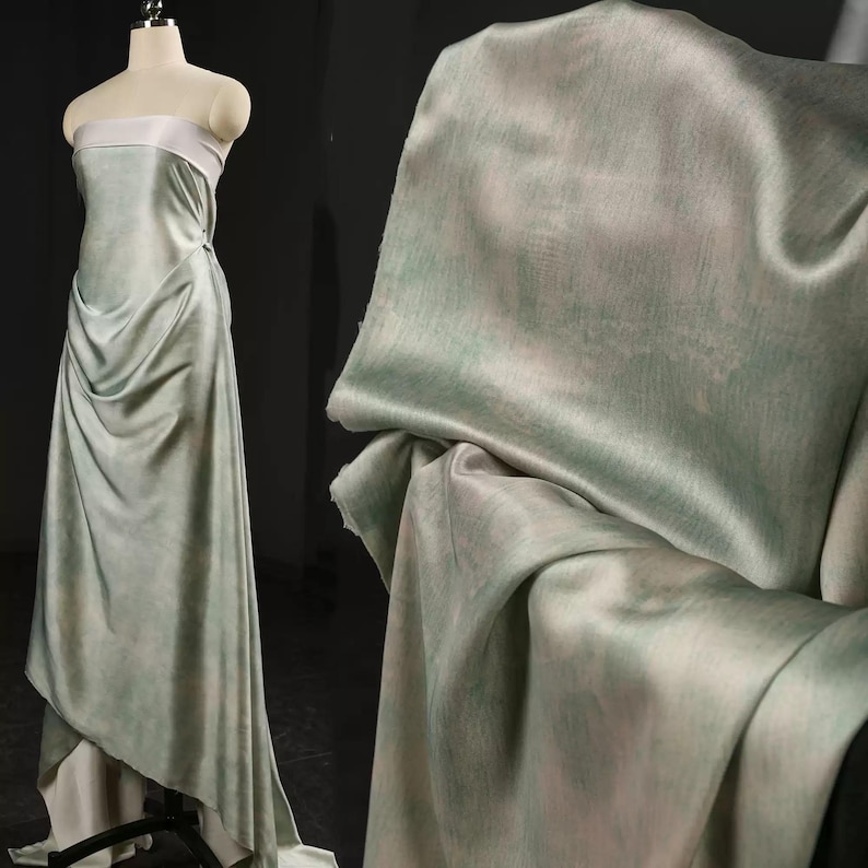 May include: A draped, floor-length gown on a mannequin, featuring a soft, flowing fabric in shades of pale green and white. The dress has a strapless bodice with a white band and a draped skirt. The fabric appears to have a subtle, watercolor-like effect.