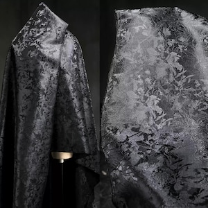 Puede incluir: Una capa negra con un estampado floral. La tela está drapeada sobre un maniquí, mostrando la textura y el diseño. La capa tiene una capucha y una silueta fluida, adecuada para disfraces o eventos especiales.