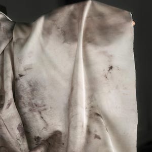 May include: A close-up of a cream-colored fabric with dark brown, irregular blotches. The fabric is draped, showing folds and textures. The material appears soft and lightweight, possibly silk or a similar textile.