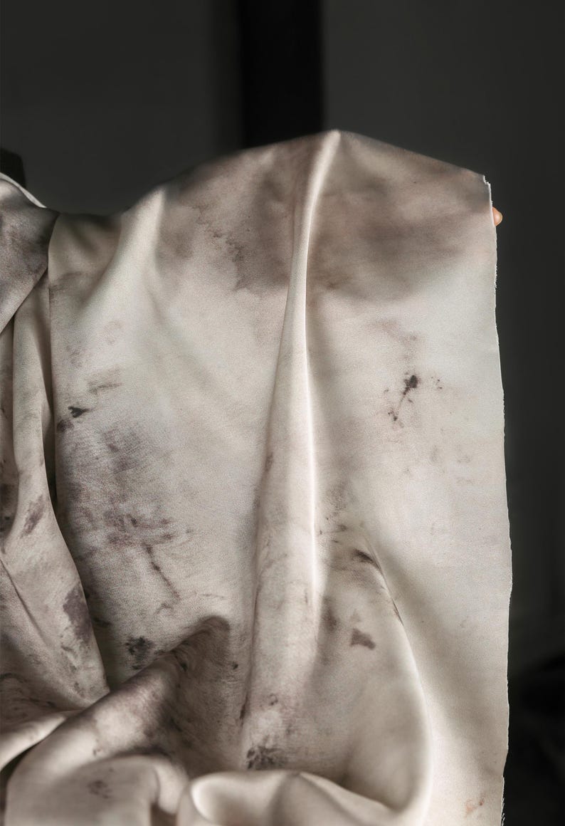 May include: A close-up of a cream-colored fabric with dark brown, irregular blotches. The fabric is draped, showing folds and textures. The material appears soft and lightweight, possibly silk or a similar textile.