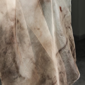 May include: A sheer, flowing fabric in shades of beige and brown, with a mottled, watercolor-like pattern. The fabric appears lightweight and delicate, with visible folds and a soft texture. The edges are finished with a subtle seam.