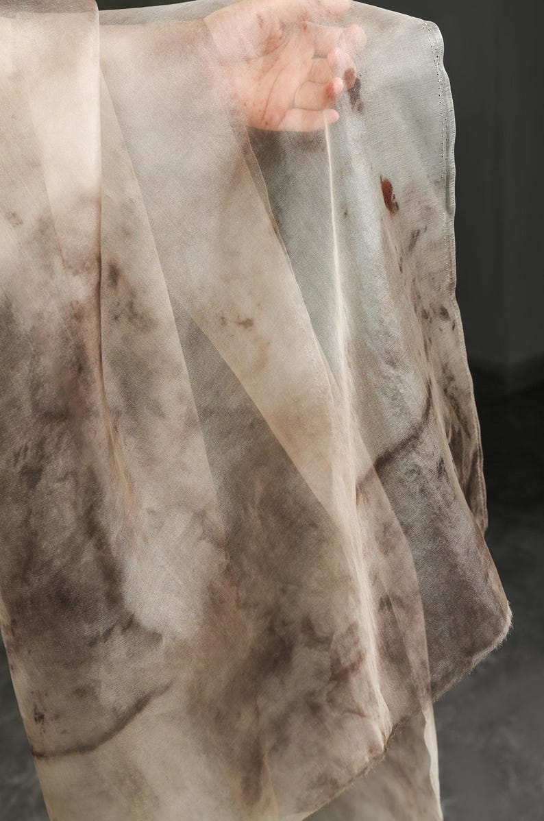 May include: A sheer, flowing fabric in shades of beige and brown, with a mottled, watercolor-like pattern. The fabric appears lightweight and delicate, with visible folds and a soft texture. The edges are finished with a subtle seam.