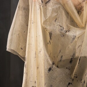 May include: A sheer, cream-colored garment with dark gray, abstract markings. The fabric appears lightweight and flowing, with a slightly textured surface. The garment has a draped appearance, suggesting it may be a shawl or wrap.