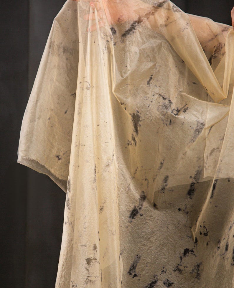 May include: A sheer, cream-colored garment with dark gray, abstract markings. The fabric appears lightweight and flowing, with a slightly textured surface. The garment has a draped appearance, suggesting it may be a shawl or wrap.