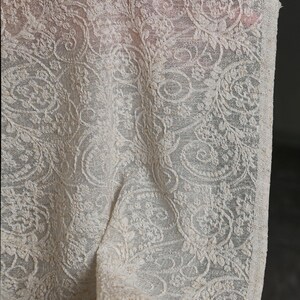 May include: Ivory-colored lace fabric with an intricate floral and swirl pattern. The delicate material is draped, showcasing the detailed design. The lace has a textured appearance, with visible details of the weave. The fabric is likely used for apparel or home decor.