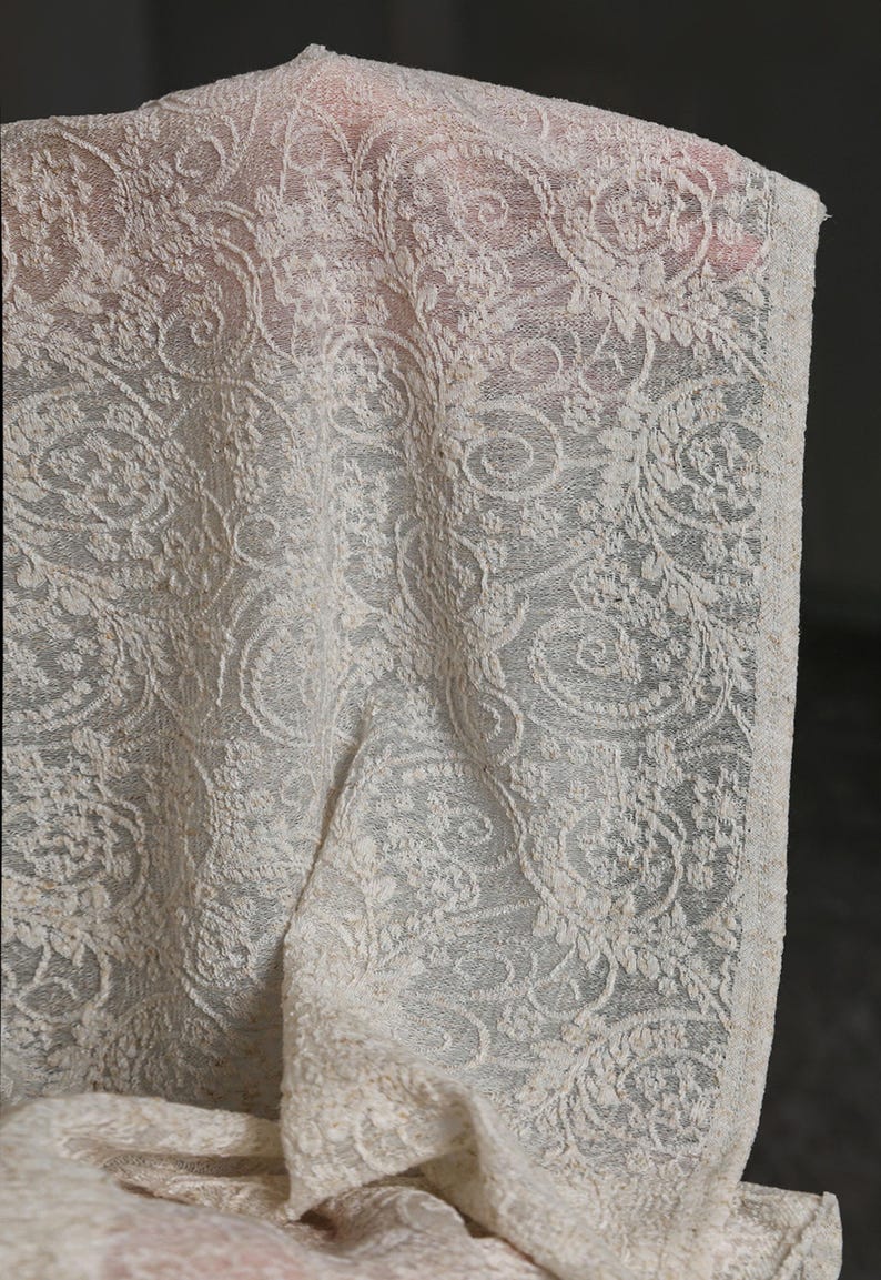 May include: Ivory-colored lace fabric with an intricate floral and swirl pattern. The delicate material is draped, showcasing the detailed design. The lace has a textured appearance, with visible details of the weave. The fabric is likely used for apparel or home decor.