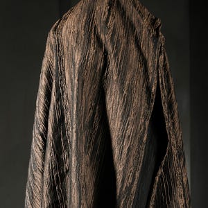 May include: A draped fabric with a textured, vertical striped pattern in shades of brown and black. The fabric is hanging, with a split visible on the right side, revealing a dark interior. The overall effect is of a rich, layered textile.
