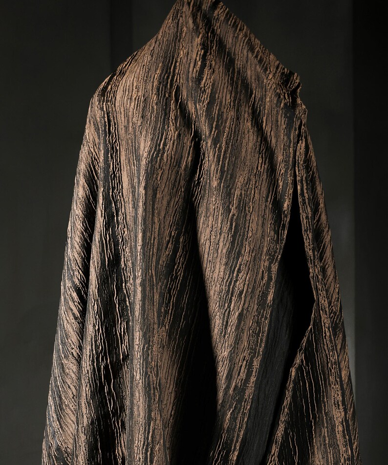 May include: A draped fabric with a textured, vertical striped pattern in shades of brown and black. The fabric is hanging, with a split visible on the right side, revealing a dark interior. The overall effect is of a rich, layered textile.