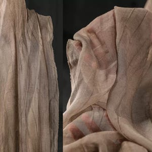 Puede incluir: Primer plano de una tela beige transparente con líneas verticales y horizontales, creando una apariencia texturizada. La tela está drapeada, con pliegues y sombras que añaden profundidad. El material parece ligero y delicado.