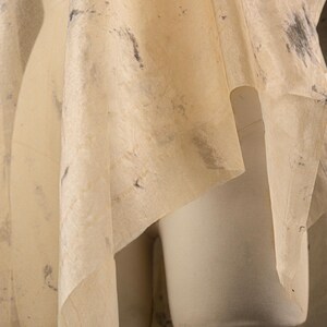 May include: A draped, cream-colored fabric with dark gray and black speckles is draped over a white dress form. The fabric has a delicate, semi-transparent quality, suggesting a textile or garment design.