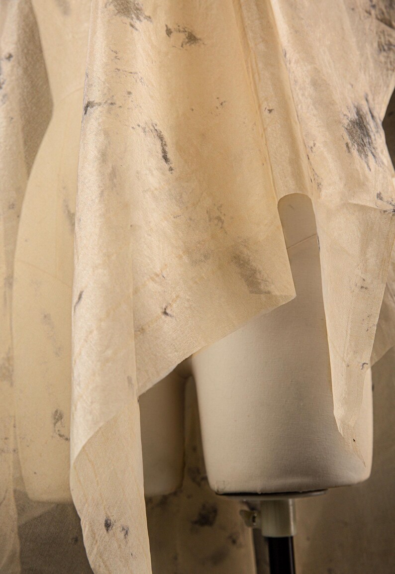May include: A draped, cream-colored fabric with dark gray and black speckles is draped over a white dress form. The fabric has a delicate, semi-transparent quality, suggesting a textile or garment design.