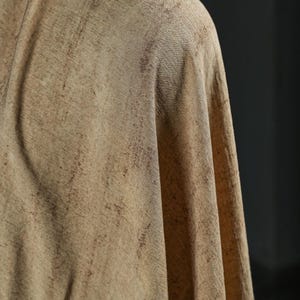 Puede incluir: Primer plano de una tela drapeada en tonos beige y marr&oacute;n. El textil tiene un aspecto texturizado con sutiles variaciones de color, creando un efecto moteado. La tela est&aacute; doblada, revelando su ca&iacute;da y textura.