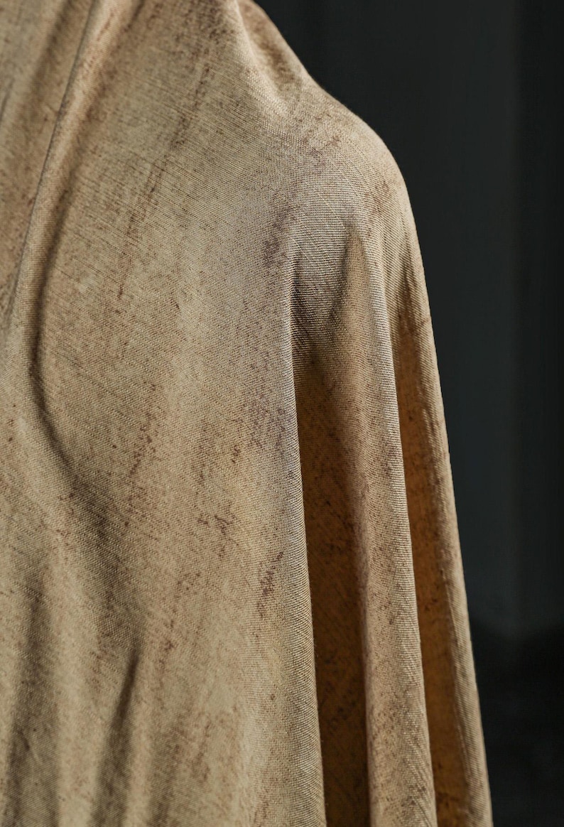 Puede incluir: Primer plano de una tela drapeada en tonos beige y marr&oacute;n. El textil tiene un aspecto texturizado con sutiles variaciones de color, creando un efecto moteado. La tela est&aacute; doblada, revelando su ca&iacute;da y textura.
