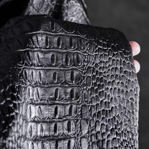 May include: Close-up of a black, textured material with a crocodile skin pattern. The material has a glossy finish and is held by a hand, showcasing the detailed scales and ridges. The overall aesthetic is dark and luxurious.