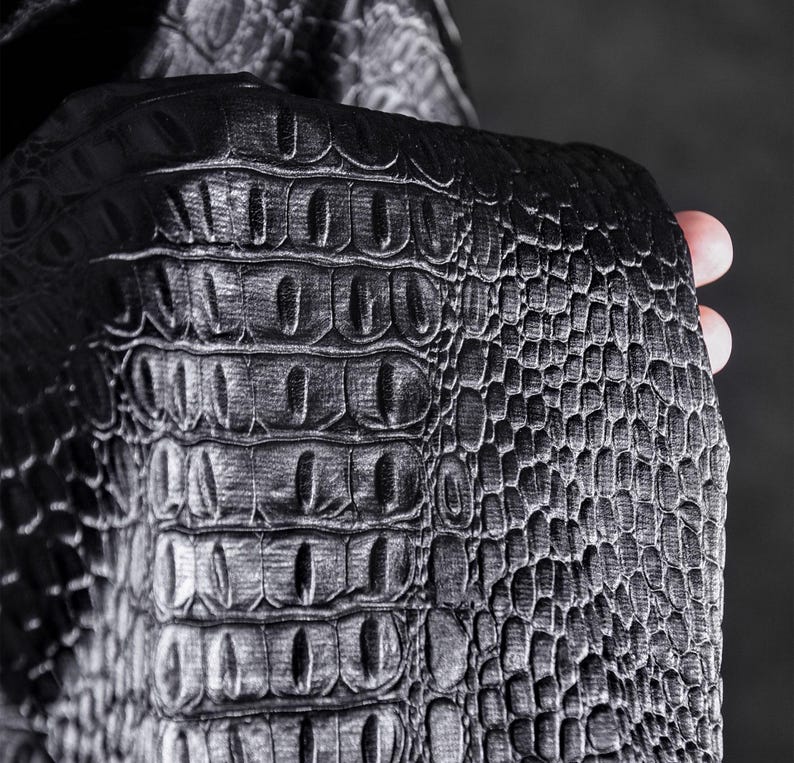 May include: Close-up of a black, textured material with a crocodile skin pattern. The material has a glossy finish and is held by a hand, showcasing the detailed scales and ridges. The overall aesthetic is dark and luxurious.